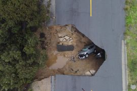An aerial view of a roadway in California that has partially collapsed, with two vehicles visible under the broken pavement