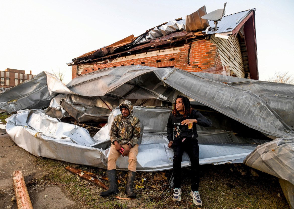 Two men sit on metal roofing in front of a broken brick home