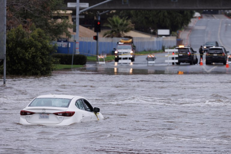 A white car sits in floodwater that reaches halfway up its doors in San Diego, California. Emergency vehicles block of the submerged road for a high point in the distance.