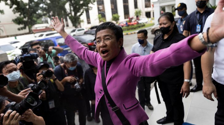 Maria Ressa wearing a purple jacket has her arms out wide outside the court, looking jubilant