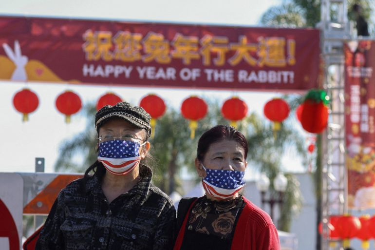 Two people stand near the location of the shooting in Monterey Park. One is wearing a cap and glasses. Both are wearing face masks showing the US flag. There is a red banner across the road behind them saying Happy Year of the Rabbit in English and Chinese.