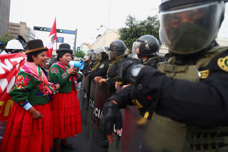 Two women in traditional Peruvian dress face ranks of riot police wearing helmets and carrying shields. One of the women is speaking through a loud hailer