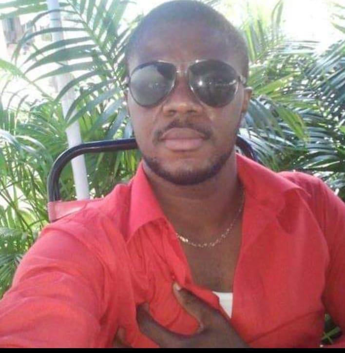 A portrait of Cletus Chimaobi Hillary. He is wearing a red shirt and aviator-style sunglasses. There are palm trees behind