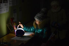 A girl at a desk drawing on a piece of paper. She has a can of pencils beside her and the desk is illuminated by a single light. Everywhere else is dark and she is wearing a winter coat and woolly hat because of the cold.