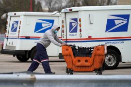 A mail carrier pushes a car through a parking lot of mail vans