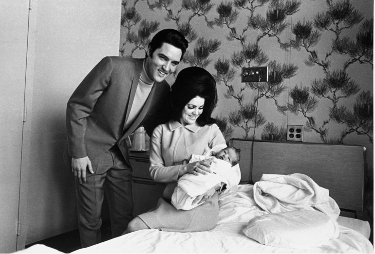 Black and white photo showing Elvis and Priscilla with newborn baby Lisa Marie in February 1968. Elvis is wearing a suit and leaning down with his arm behind Priscilla who sits on the bed cradling Lisa Marie and smiling at her 