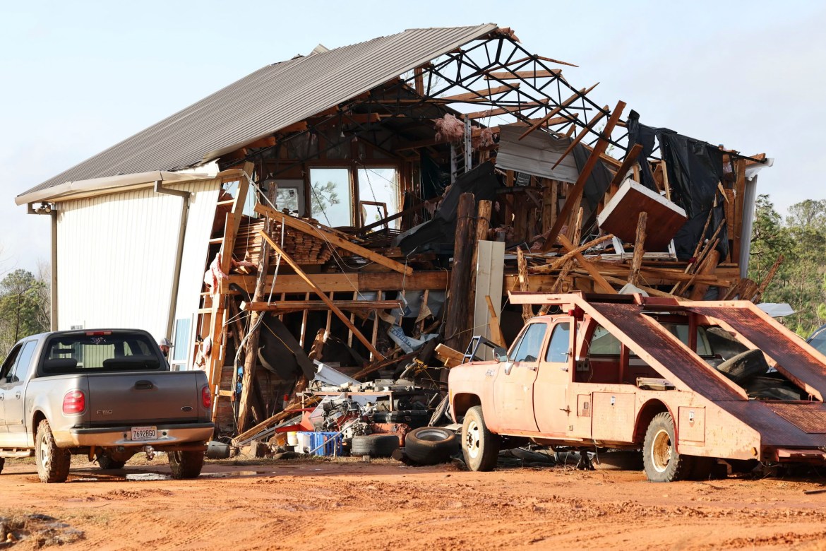 A home with a ripped-up roof and no front wall, with debris scattered on surrounding vehicles