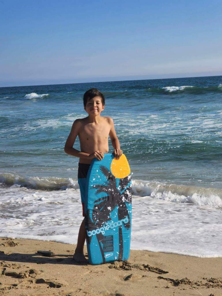 Honor Beauvais on vacation at a California beach, wearing swim trunks and carrying a boogie board.