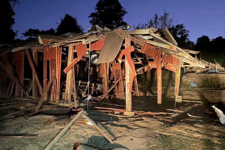 The ruins of a village school. Some wooden boards are still standing, but the wooden structure has generally collapsed. The sky is a deep purpose. It looks to be nighttime.