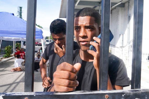 A Cuban man calls his grandmother from a US border facility. He is standing behind bars and holding mobile phone to his ear while clutching a bar. A man stands behind him, looking down with his hand to his mouth.