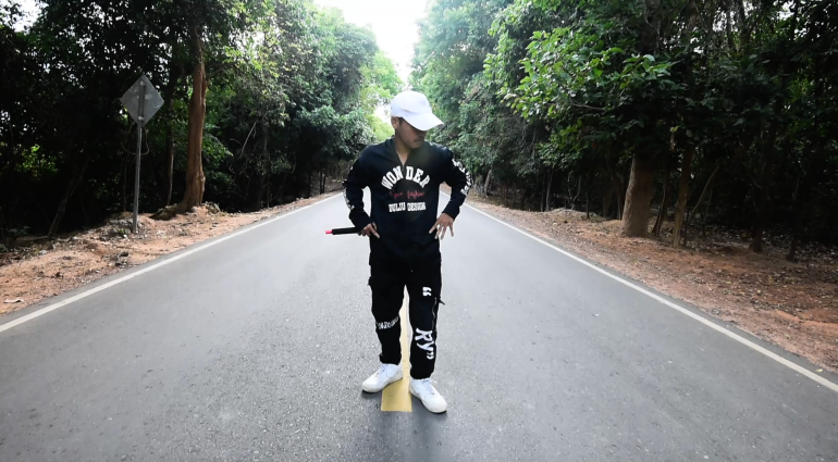 Kea Sokun in black sweat pants and sweat shirt with the word WONDER written across the chest. He is performing in a rap video. He is standing on a road and there are trees running down each side. He has his hands on his hips and is s holding a microphone in his right hand and looking down and away from the camera to his left. He's wearing a white baseball cap. His trainers are also white.