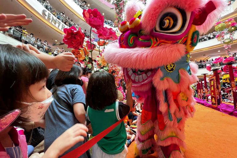 Children gather around a lion after a performance in Malaysia to get mandarin oranges. The lion has big eyes rimmed in pink fur . Its ears and top lip are also covered in pink fur. The children look excited. There are lots of people looking down on the concourse from higher levels.