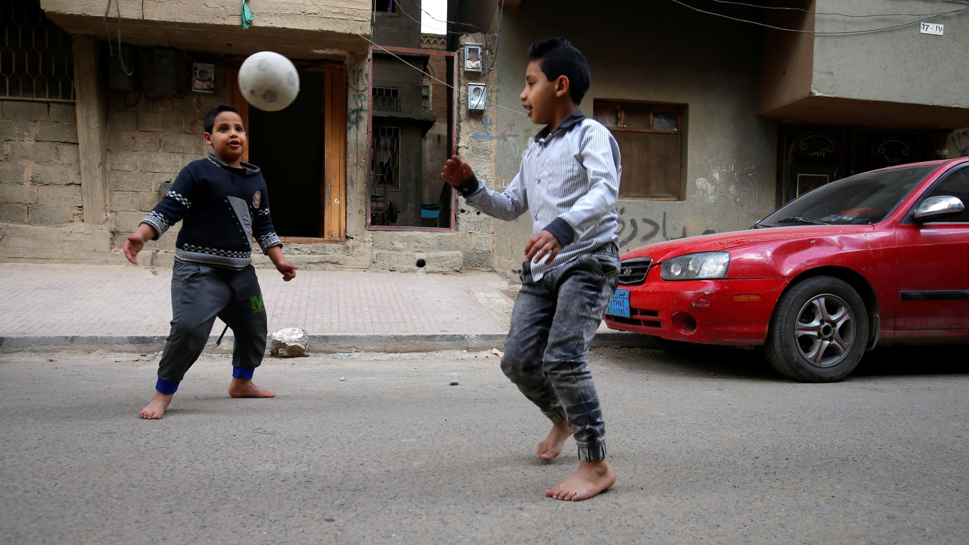 A photo of two kids playing football in the street, outside of a building with a red car in front of it.