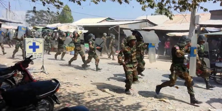 Police in riot gear chasing after garment workers protesting for higher wages during the 2014 Veng Sreng protests in a still from the Workers Blood video. It is dusty and hot. The police are wearing fatigues and holding their plastic shields in front of them. They have large truncheons in their hands