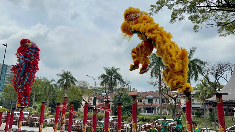 Lion dancers in full costume leap from one high pole to another. The leaping lion is yellow and furry. There is a second lion in front which is red and blue and the two performers who make up the lion are standing on a single pole. The man who makes up the rear of the lion is holding up the person who is its head and front legs. The performance is outside and there are houses and trees behind.