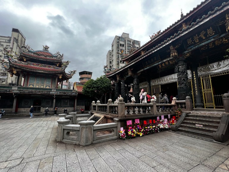 A view of the Longshan temple in Taiwan. It has an ornate roof and there are steps leading up to the prayer halls. People are queuing up behind a balustrade in the centre of the photo to get into the temple. Flowers have been placed around the bottom.