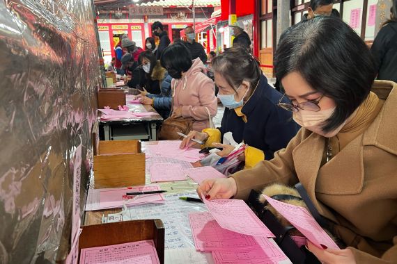 A woman in coat and face mask fills out her particulars so the gods at the Longshan temple will know her. The form is pink and there are lots other people filling it out as well