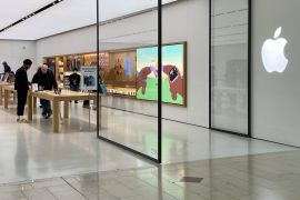 A view through the open glass doors of an Apple store. There are a number of people inside looking at the products. There is a big Apple logo on the wall