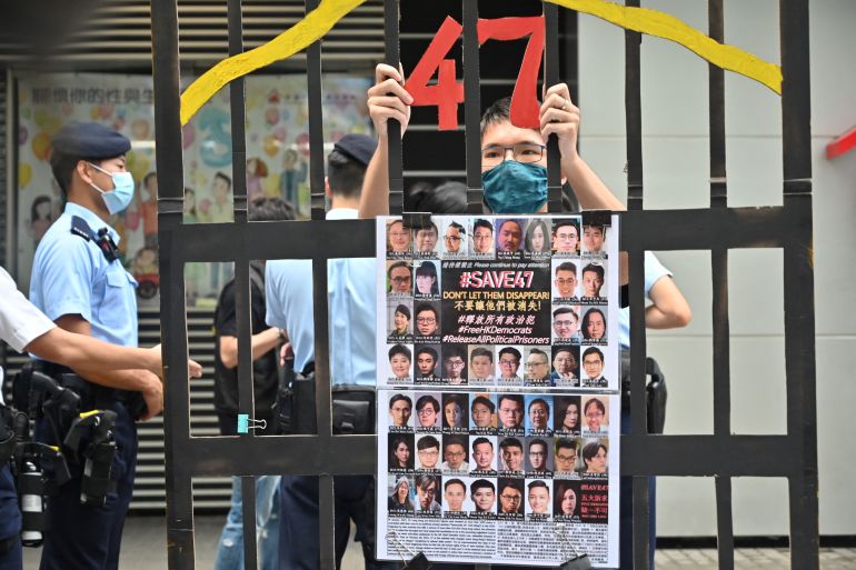 A man stands behind a mock-up of the bars of a jail cell holding a banner showing the 47 Hong Kong activists and legislators charged under security laws for arranging their own primary. The number 47 is in red at the top of the bars. There are masked police officers behind