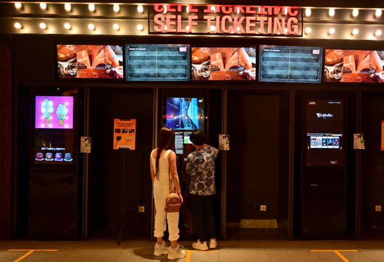 Two people buying cinema tickets in Jakarta. There are TV screens above them and the words SELF TICKETING is lit up