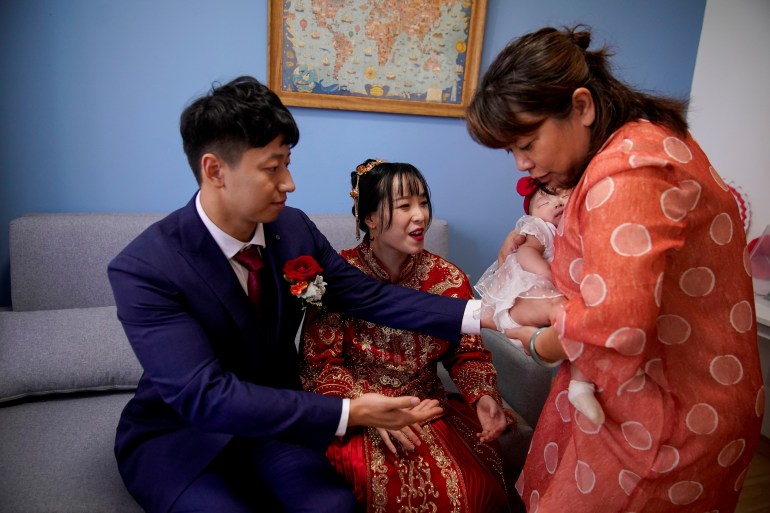 A married couple with their young baby. The bride is wearing a traditional red outfit and the groom is in a Western-style suit. An aunt is holding their baby 
