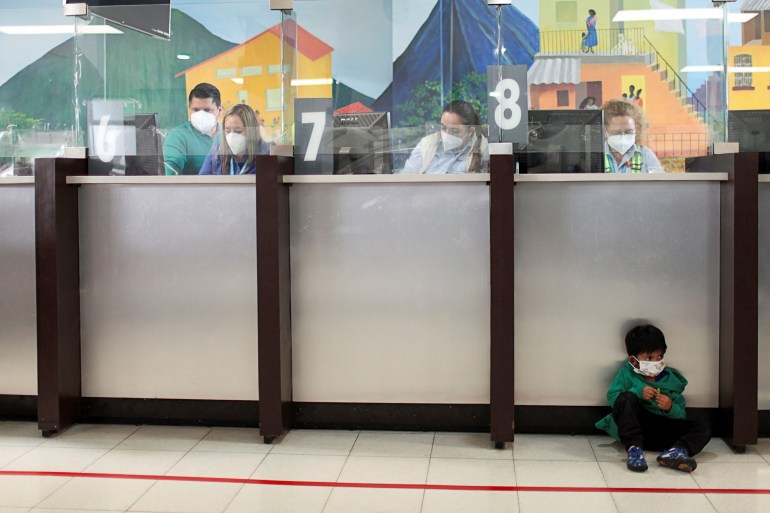 A boy sits on the floor, while behind him, adults look on computers behind a glass barricade