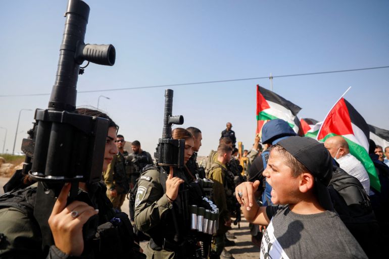 Palestinian boy standing in front of an armed Israeli soldier during protest. Behind him are other unarmed protesters with Palestinian flags coming up to a line of armed Israeli soldiers.