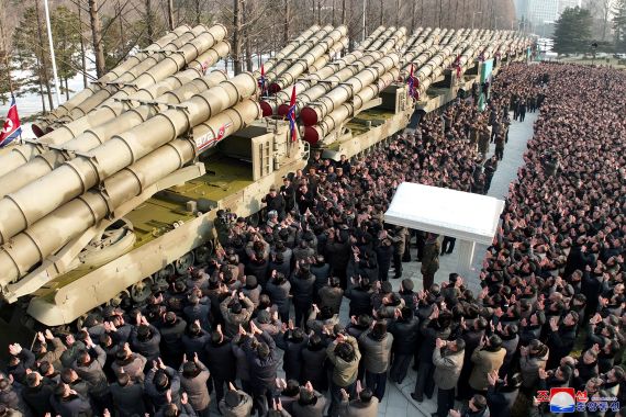 Super-large weapons launchers on display in North Korea. There are lots of men crowded around clapping. Flags are positioned on the weapons. Kim Jong Un is standing near one of the launchers, his right hand raised. He is being filmed.