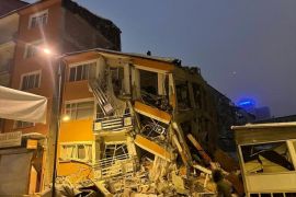 A collapsed building in Malatya, Turkey. The building is illuminated. A man is walking past.