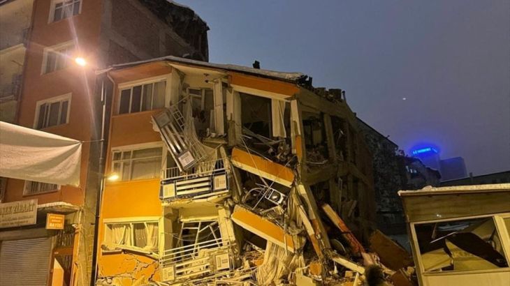 A collapsed building in Malatya, Turkey. The building is illuminated. A man is walking past.