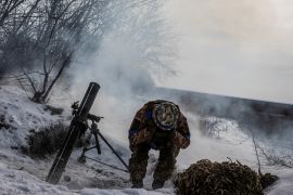 A soldier crouches and covers his ears with both hands as a mortar launcher fires next to him. There is snow everywhere and the landscape is grey