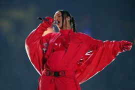 Rihanna singing on the Super Bowl stage. She is wearing a red outfit with sweeping sleeves.