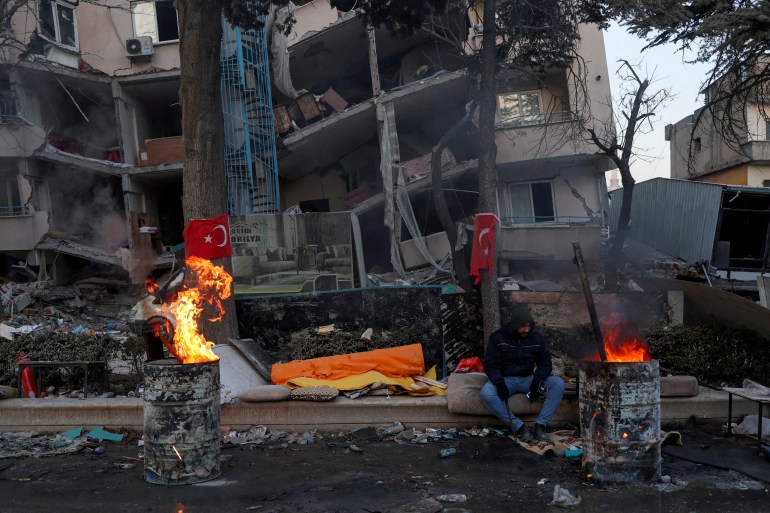 A man keeps warm by a fire in front of a damaged building in the aftermath of a deadly earthquake in Kahramanmaras