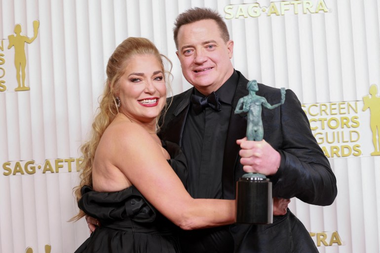 Brendan Fraser holdling his best actor award for The Whale. He is dressed in black and looks happy. He is with his parter Jeanne Moore who is holding him around the waist.