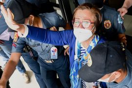 Leila de Lima waving to supporters during a court appearance in January. She is wearing a blue and white stripy top with a blue cardigan and scarf around her neck. She is also wearing glasses and a face mask.