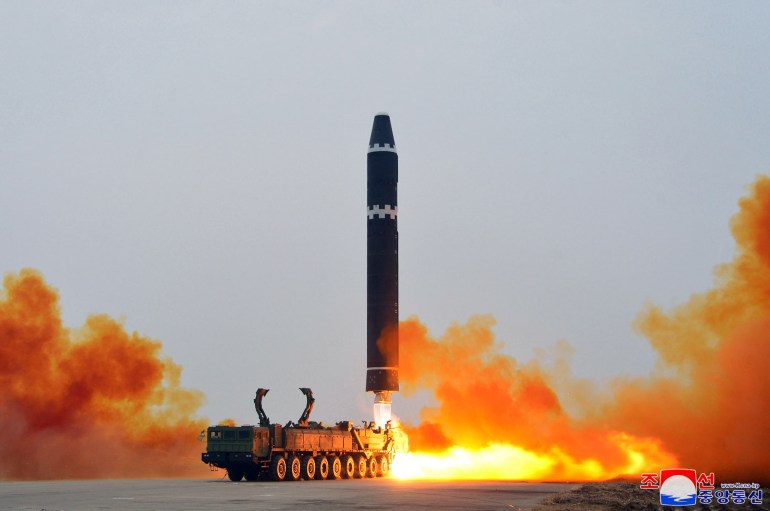 A test firing of the Hwasong-15 in a still from KCNA. The missile is black with a white checkered pattern close the cone. It is lifting vertically into the air in clouds of orange smoke. There is a transport vehicle to one side.