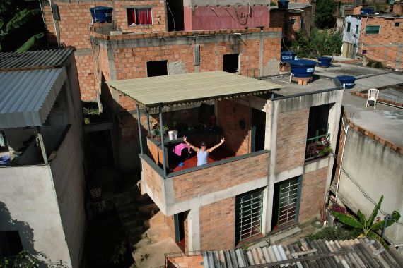 An aerial view showing Brazilian musical artist Kdu dos Anjos gesturing at his house, which was designated "Building of the Year 2023" by the specialized reference site ArchDaily, in Aglomerado da Serra, a favela complex on the outskirts of Belo Horizonte, Minas Gerais State, Brazil. A person on a balcony of the house is looking towards the camera with arms outstretched.