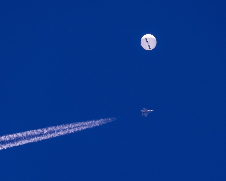 A white balloon against a bright blue sky with a fighter jet and its trails beneath it.