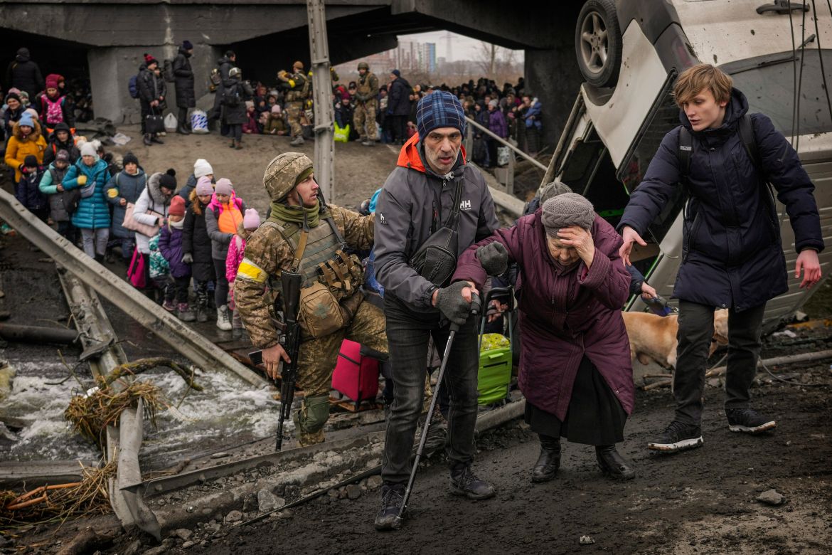 An elderly woman is assisted while crossing the Irpin River on an improvised path under a bridge that was destroyed by Ukrainian troops designed to slow any Russian military advance, while fleeing the town of Irpin