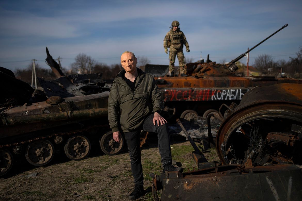 A civilian wears a Vladimir Putin mask as a spoof, while a Ukrainian soldier stands atop a destroyed Russian tank in Bucha