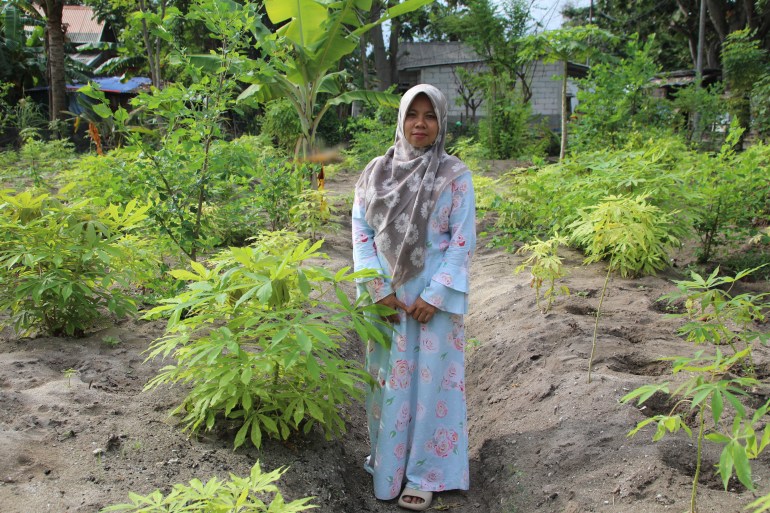 Asmania standing in the vegetable garden she and some other women have set up as an alternative source of income. She is wearing a pale blue top and long skirt with a floral headscarf