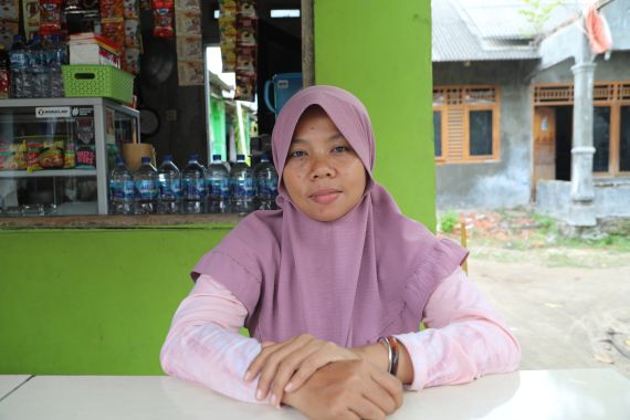 A portrait of Asmania. She is sitting at a table outside a cafe painted green. She is wearing a pale pink top and mauve headscarf. She has her hands resting on the table in front of her and looks relaxed