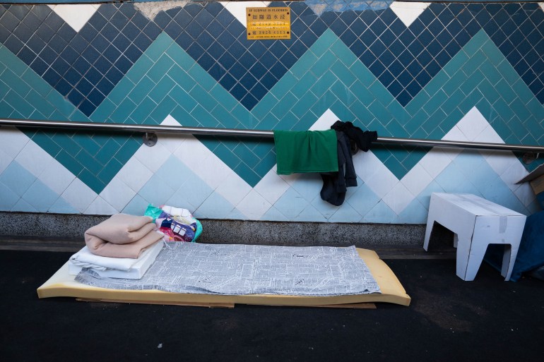 A thin foam mattress with blankets folded neatly on top at one end in an underpass in Hong Kong. The wall behind is tiled in blue, white and teal chevron and there is a handrail. Some clothes have been hung on it.