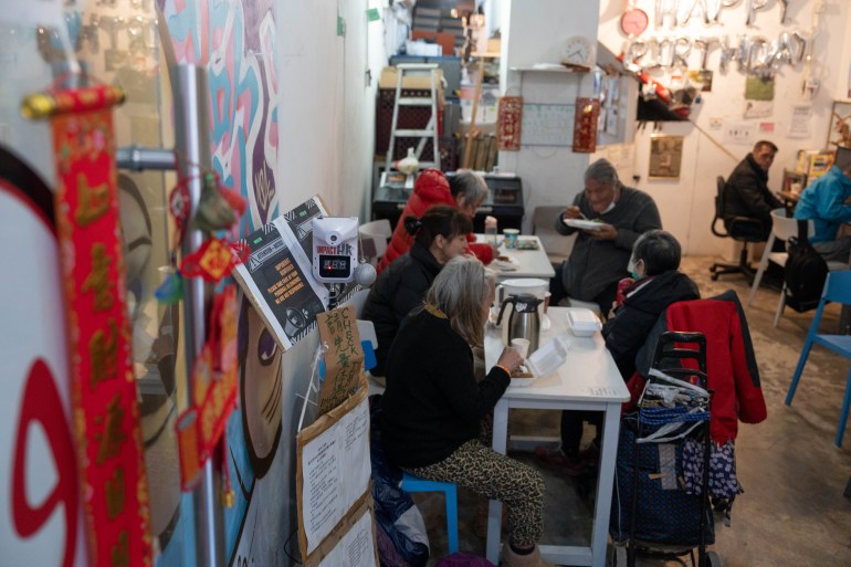 People eating inside the ImpactHK centre. They are seated at tables with their food and some are drinking tea and coffee. They are dressed in winter clothes. There are notices in Chinese and a red banner on the walls and door.