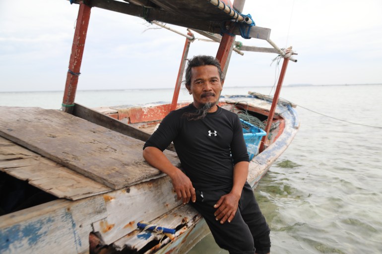 Bobi leaning against a fishing boat which is in shallow water. He is wearing black trousers and a black t-shirt