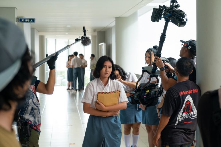 A scene from the film Dear David. A schoolgirl standing in a corridor hugging some files to her chest. She looks worried. There are other girls looking at her and a group of students behind. The camera crew is filming the scene