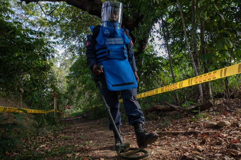 A man with a blue blast vest that reads, in Spanish, "humanitarian de-mining" sweeps the ground with a metal detector. Yellow caution tape forms a barrier behind him.
