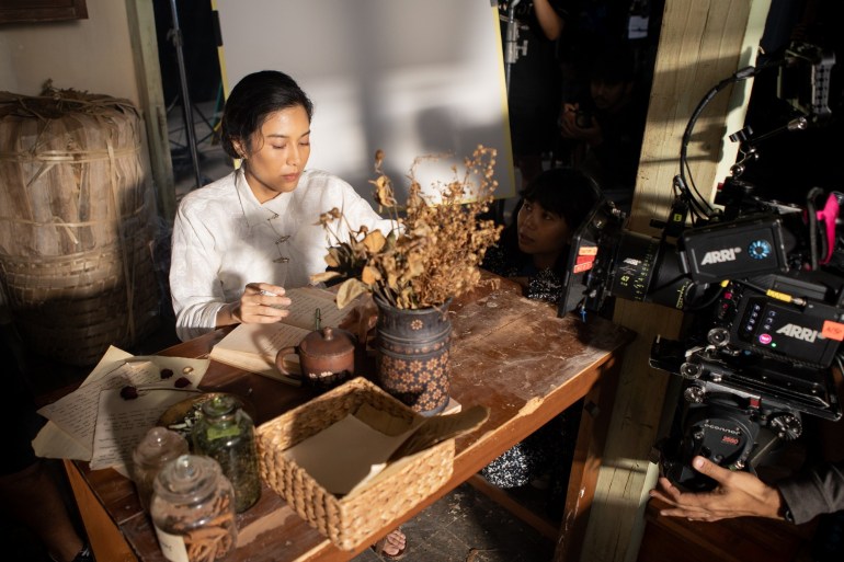 A scene from the period drama Gadis Kretek. A woman is sitting at a desk. She is wearing a white top and a note book is open in front of her. There are also sheets of paper, a vase of dried leaves, and other glass jars. The desk is dusty. The scene is being filmed and a member of the crew is squatted down next to the desk
