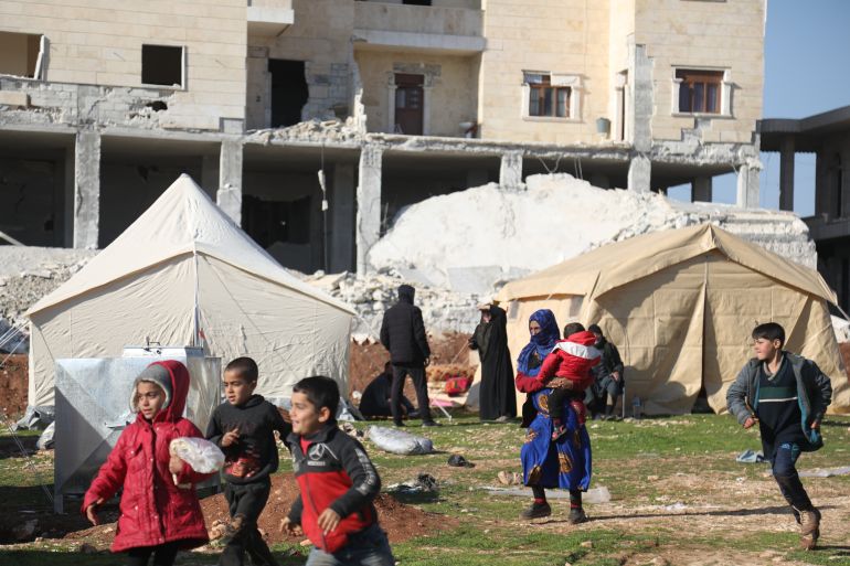 children run and play in front of shelter tents set up in front of destroyed buildings