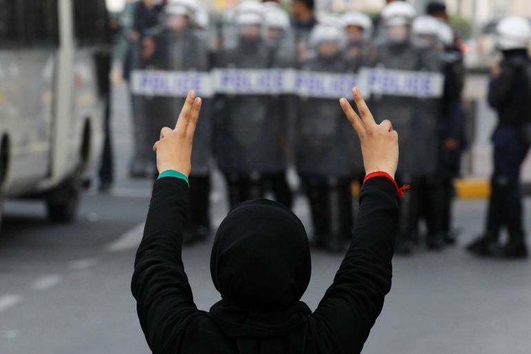 A woman holding up her fingers in a V for Victory sign towards a row of riot police in Manama, Bahrain. She has her back to the camera, is wearing black and has red and green ties around each wrist